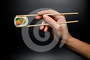 hand holds piece of Philadelphia sushi roll with smoked salmon, avocado, cucumber and green calve with bamboo chopsticks