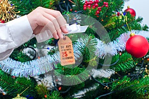 A hand holds a tag with a bow for a gift `do not open until December 25th` on the background of the Christmas tree