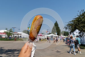 Hand holds up a pronto pup smothered in mustard at the fairgrounds in summer