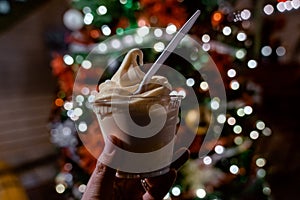 Hand holds up a soft serve pineapple vanilla swirl ice cream in a cup, with Christmas tree bokeh in background, at night