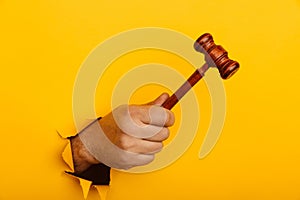 Hand holds a wooden gavel through torn yellow wall. Law or auction concept