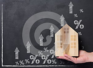 A hand holds a wooden house against the background of a graph with growing indicators, the concept of rising house prices