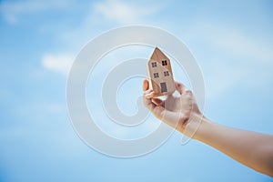 Hand holds wooden house model on blue sky with clouds background. Concept saving for building dream home