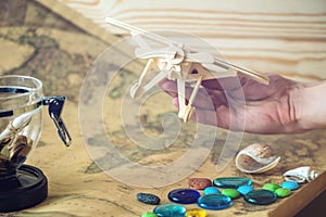 Hand holds wooden toy plane on a world map with colored stones and shells from the sea in a retro style