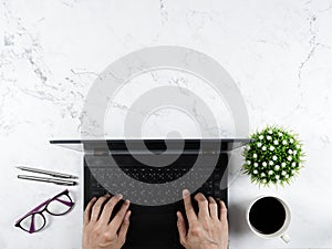 Hand on keypad laptop with glasses silver pen coffee cup flower vase on marble table workplace concept copy space top view