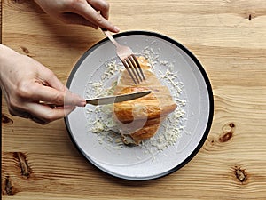 Hand with knife and fork above the croissant sandwich filled with lettuce leaf, tomato, and white cheese on a white plate with