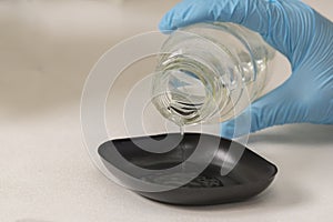 hand of a lab technician with blue nitril gloves pouring liquid into a black plastic container in a scientific laboratory.