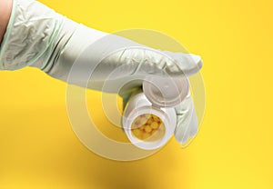 Hand in latex glove holding container of pills on bright yellow background