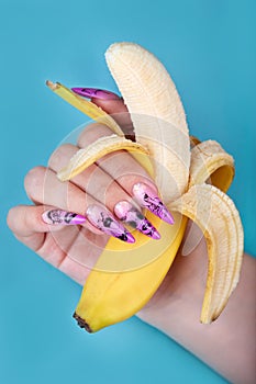 Hand with long artificial manicured nails colored with pink nail polish with summer design holding a banana