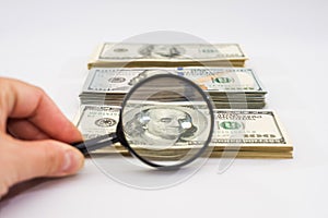 Hand with a magnifying glass checks one hundred dollar bills against a white