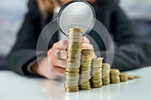 Hand with magnifying glass and money coins stack on office desk.