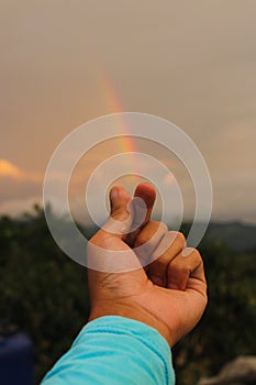 Hand making a love sign with rainbow background on a cloudy sky after rain in the afternoon.