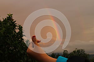 Hand making a love sign with rainbow background on a cloudy sky after rain in the afternoon.