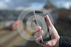 Hand of a man with a mobile phone, station, railroad tracks and train blurred in the background, concept for tracking and ticket