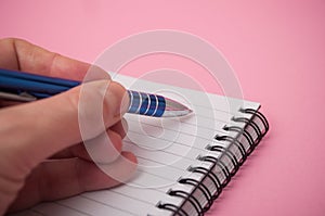 hand of man writing with blue pen on spirales note book on pink background