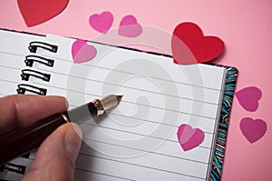 hand of man writing with vintage pen on spirales note book with paper hearts on pink background