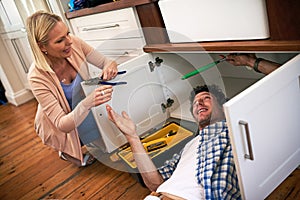 Hand me those pliers, would you darling. a man fixing pipes under his kitchen sink while his wife watches.