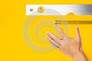 Hand, measure tape and aluminium spirit level tool on yellow background. Construction fluid level with air bubble, flat lay.