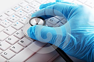 Hand with medical glove holding a stethoscope on computer keyboard. Computer repair service or health diagnosis concept