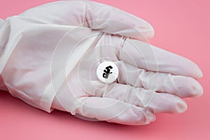 A hand in a medical glove holds a pill with a riel sign on a pink background. Expensive medicine in Cambodia. Pharmaceutical