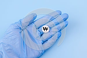 A hand in a medical glove holds a pill with a won sign on a blue background. Expensive medicine in Korea