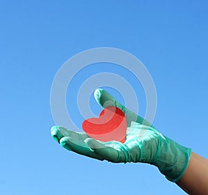 Hand in a medical glove holds a red heart on blue sky background