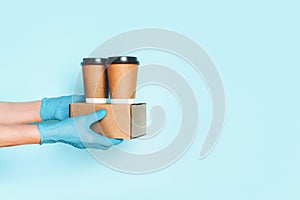 Hand in medical gloves carrying paper coffee cup coffee, lunch box on blue background. Banner, copy space. Contactless delivery
