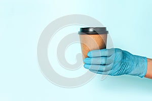 Hand in medical gloves carrying paper cup of takeaway coffee on blue background. Banner with copy space. Contactless delivery