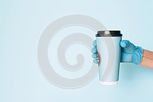 Hand in medical gloves carrying paper cup of takeaway coffee on blue background. Banner with copy space. Contactless delivery