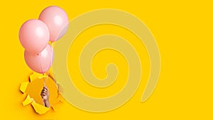 Hand of millennial african american lady holding three pink balloons through hole in yellow paper, panorama