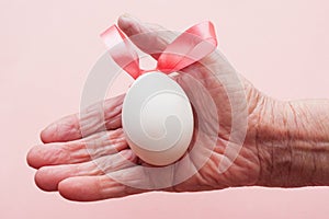 Hand of an old woman holding white egg with pink bunny-shaped ribbon.