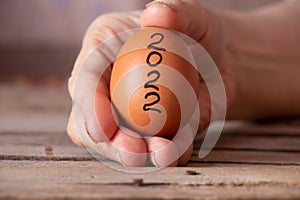 The hand of an old woman holds in her hand a chicken egg with the text 2022 on a wooden table in the kitchen, a chicken egg photo