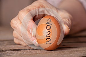 The hand of an old woman holds in her hand a chicken egg with the text 2022 on a wooden table in the kitchen, a chicken egg photo