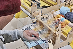 Hand of older man, who weaving small rug with pattern on wooden manual table loom.