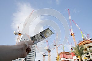 Hand with one hundred american dollars banknotes on the background of a cranes and building construction. Construction industry.