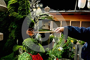 Hand of people in jacket holding a Japanese dipper scoop and pouring water on head of Buddha statue covered by lichens.