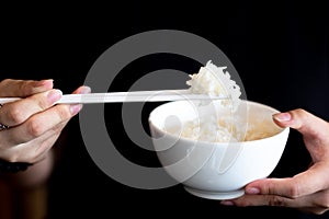 Hand using white chopsticks holding hot steamed rice in white bowl on dark background