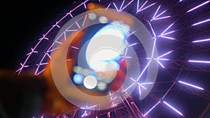 Hand with phone taking photo. Ferris wheel illuminated at night.
