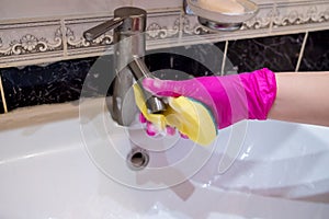 A hand in a pink glove washes a washbasin in the bathroom with a sponge with detergent