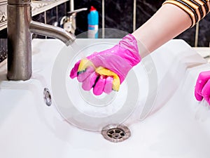 A hand in a pink glove washes a washbasin in the bathroom with a sponge with detergent