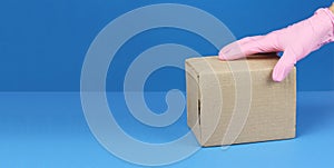 Hand in pink medical glove hold cardboard box on table, goods delivery in virus quarantine, copy space blue background