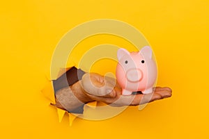 Hand with a pink piggy bank through a yellow paper hole. Savings and investment concept
