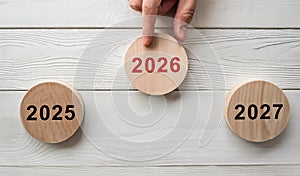 Hand Pointing to 2026 Wooden Disc Between Years 2025 and 2027 on White Table