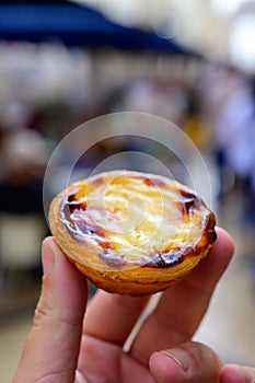 Hand with Portugal`s traditional sweet dessert Pastel de nata egg custard tart pastry and view on street in Lisbon, Portugal