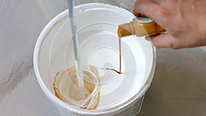 Hand pouring brown liquid in bucket with white paint.