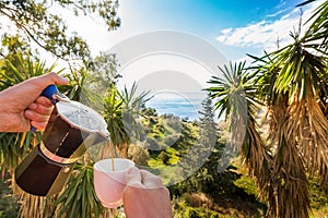 Hand Pouring Coffee In Cup against ocean, blue sky and exotic trees