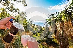 Hand Pouring Coffee In Cup against ocean, blue sky and exotic trees photo