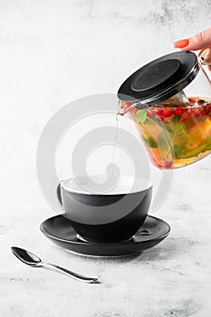 Hand pouring cranberry, orange and mint or yellow tea from glass teapoon into black cup isolated on marble background. Overhead