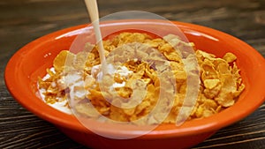 Hand pouring milk from a big bottle into the bowl full of cornflakes on wooden table. Slow motion. Side view. Close up