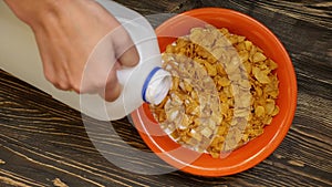 Hand pouring milk from a big bottle into the bowl full of cornflakes on wooden table. Slow motion. Top view. Close up